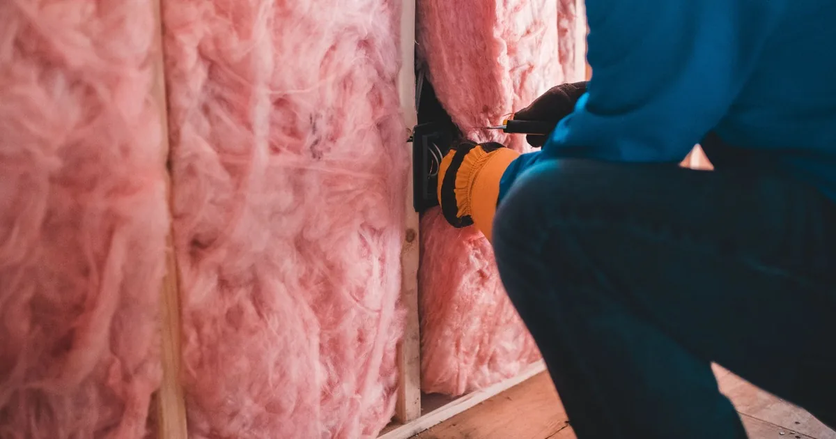 Gloved worker fitting pink fibrous insulation between wood framing