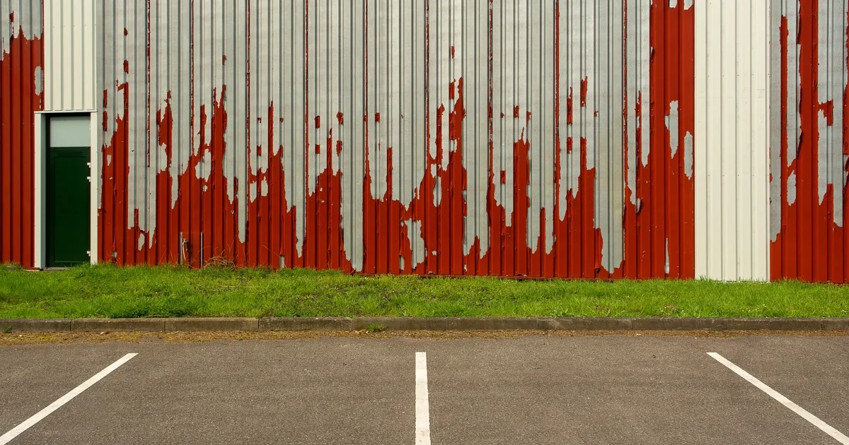 Peeling red coating on corrugated metal wall panels beside an empty parking lot